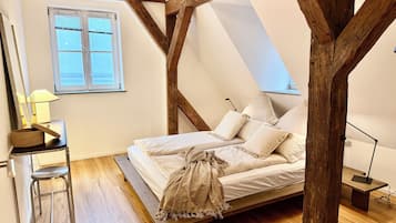 Ein Schlafzimmer mit geneigter Decke, Holzbalken, einem Bett mit weißer Bettwäsche, einem Schreibtisch mit Lampe und einem Fenster mit Jalousien.