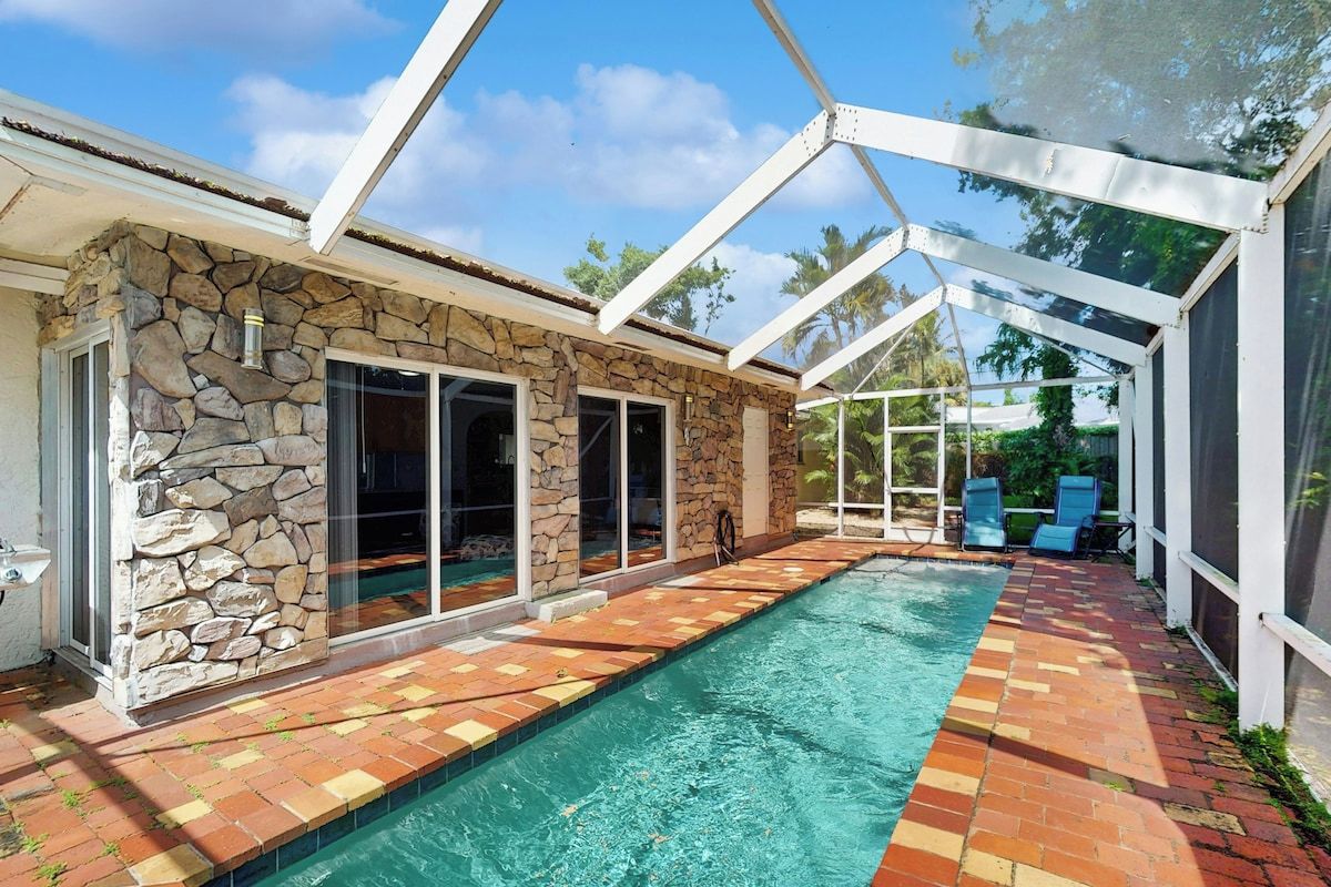 A swimming pool with a stone wall and glass doors, surrounded by a covered patio area.