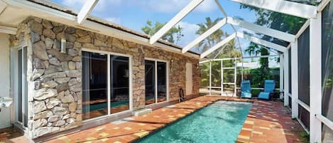 A swimming pool with a stone wall and glass doors, surrounded by a covered patio area.