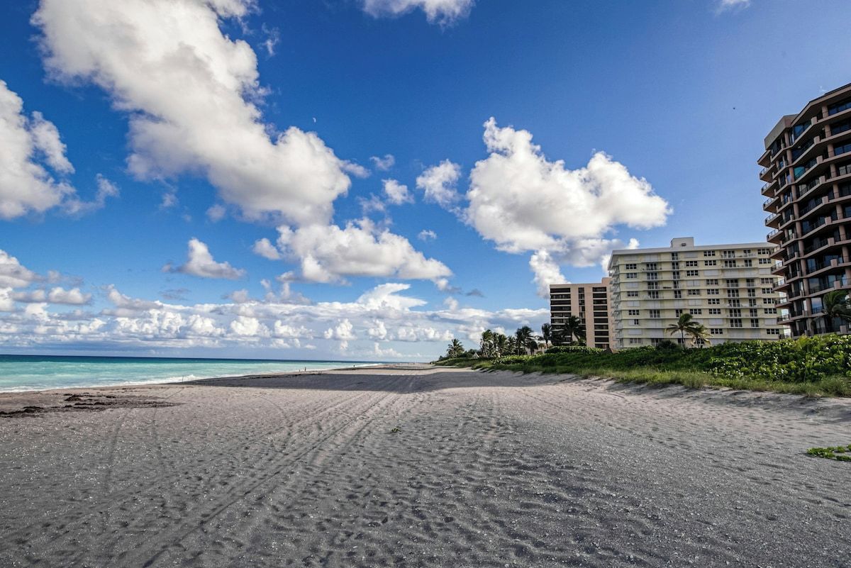 Ein Strand mit blauem Himmel, sandigem Ufer und einem mehrstöckigen Gebäude mit Balkonen.