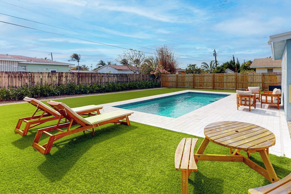 Un jardin avec une piscine, des chaises longues en bois, une table en bois et une clôture blanche.
