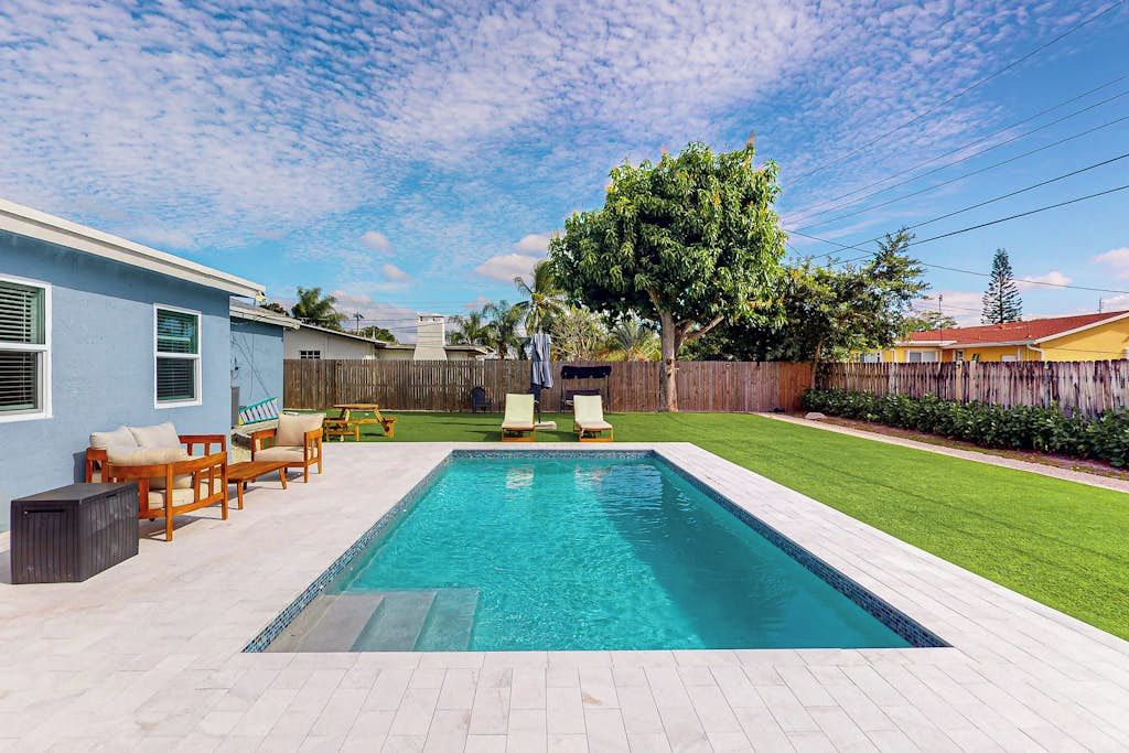 A rectangular swimming pool with a tiled deck, surrounded by a wooden fence, and a grassy lawn with a tree.