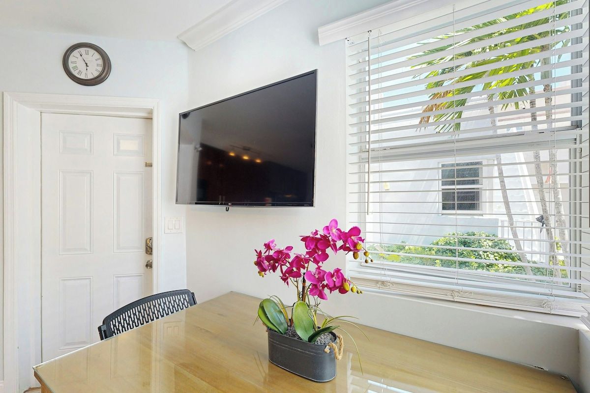 A dining area with a wooden table, a black chair, a potted orchid plant, a wall-mounted flat-screen TV, and a window with blinds.