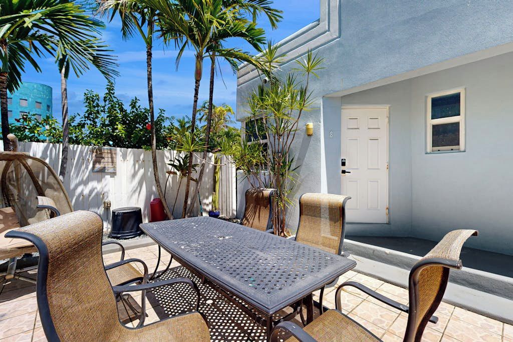 An outdoor patio area with a table and chairs, a white door, and a view of palm trees and a building in the background.