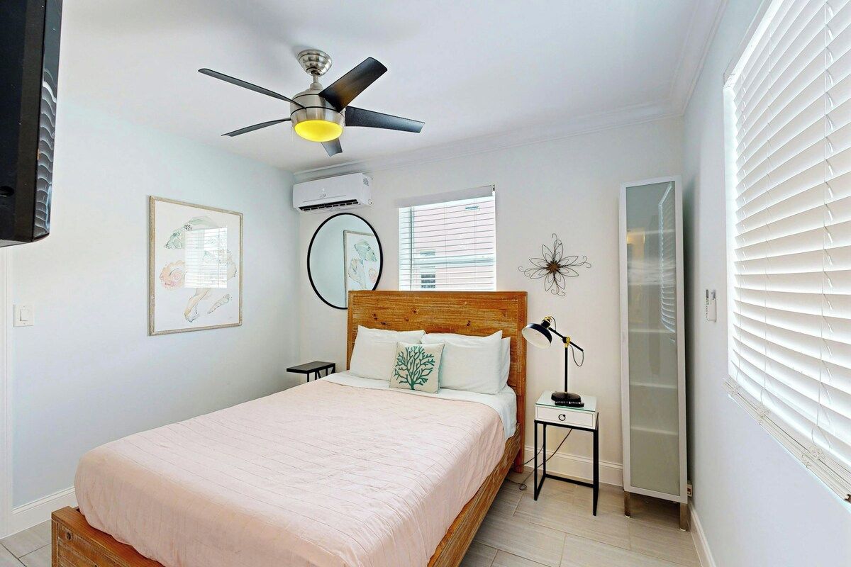 A bedroom with a bed, a ceiling fan, an air conditioning unit, a bedside table with a lamp, and a window with blinds.