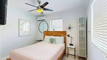A bedroom with a bed, a ceiling fan, an air conditioning unit, a bedside table with a lamp, and a window with blinds.