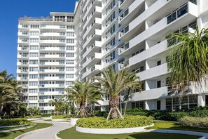 Hotel com vários andares, design arquitetônico moderno, com varandas e um jardim arborizado com palmeiras.