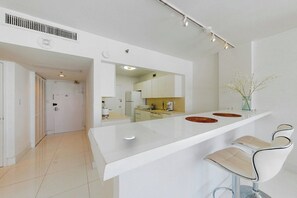 Eine moderne Küche mit weißer Arbeitsplatte, zwei Barhocker und Blick in den angrenzenden Essbereich.