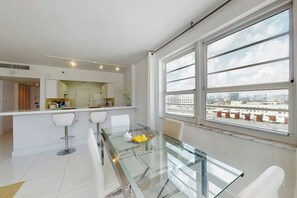 Cozinha moderna com armários brancos, uma ilha central e uma área de jantar com mesa de vidro e cadeiras brancas.
