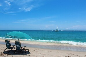 Ein Strand mit zwei Stühlen unter einem türkisblauen Sonnenschirm, ein Segelboot in der Ferne und ein klarer, blauer Himmel.