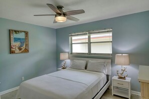 Ein Schlafzimmer mit einem Bett, Nachttischen, einem Deckenventilator, einem Fenster mit Jalousien und einem Wandbild.