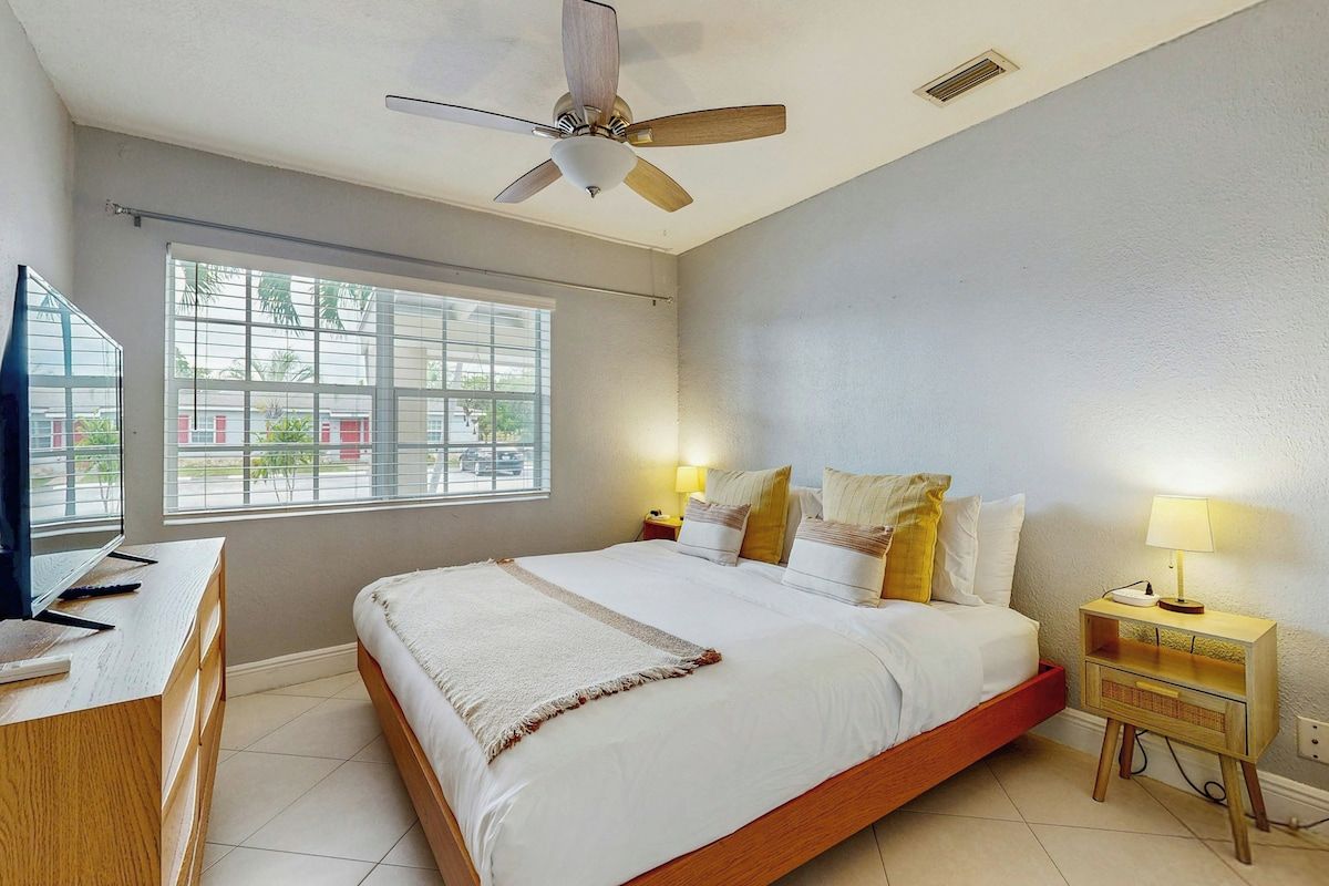Quarto com cama, luminárias de cabeceira, ventilador de teto, janela e escrivaninha com computador.