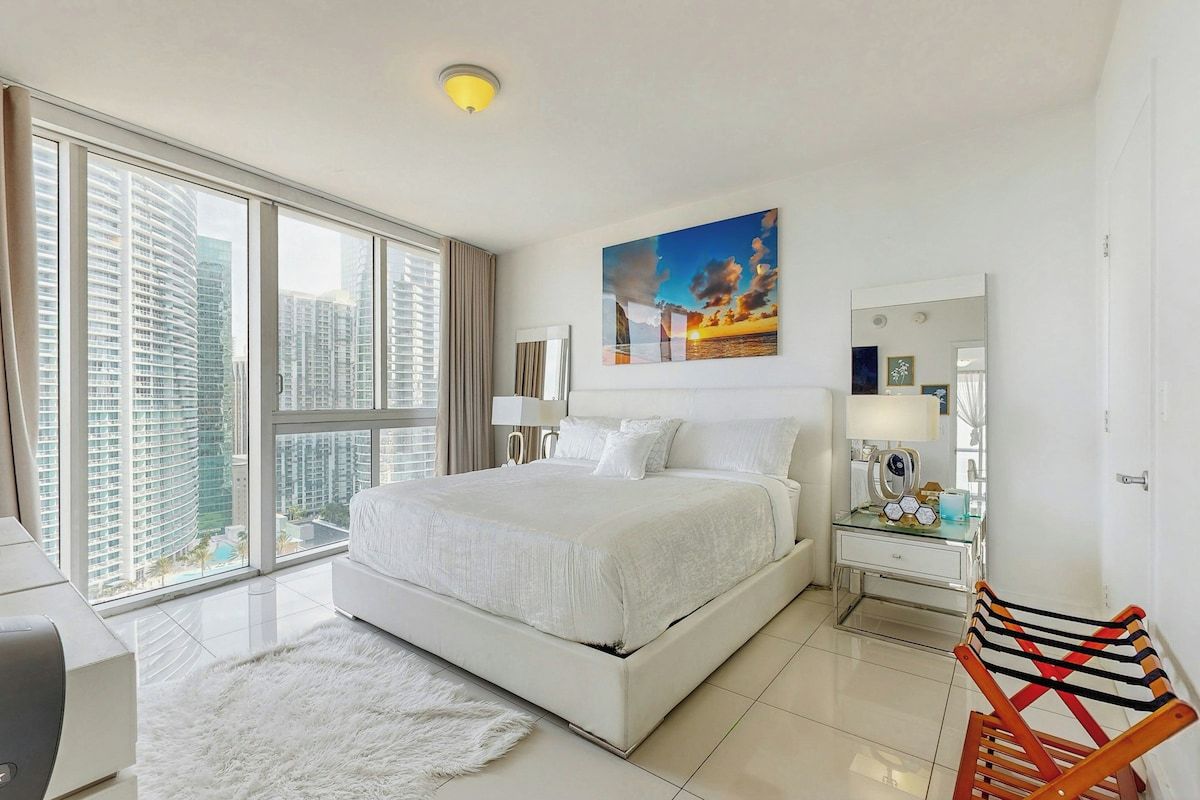 A modern hotel room with a large bed, a desk, a chair, a large window with a city view, and a painting on the wall.