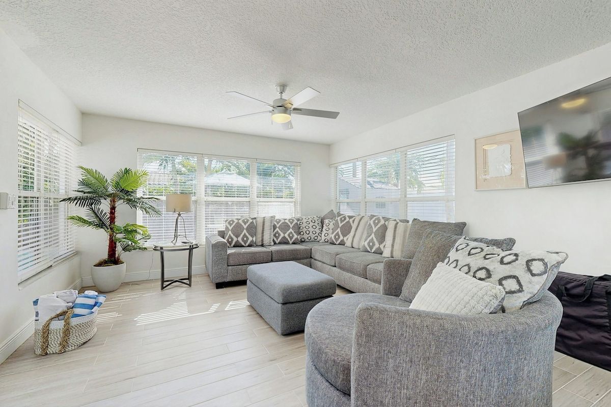 A living room with a sectional sofa, ottoman, ceiling fan, potted plant, and a framed picture on the wall.