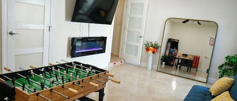Een kamer met een tafelvoetbalspel, een aan de muur bevestigde televisie en een bank met kussens.