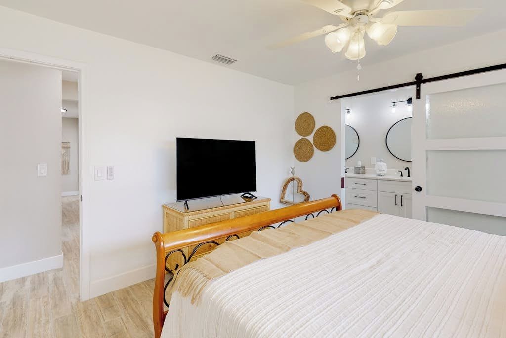 Un dormitorio con cama, televisor, ventilador de techo y baño con espejo y lavamanos.