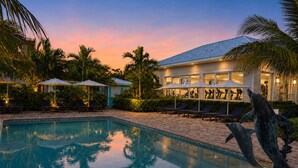 A swimming pool with lounge chairs and umbrellas, surrounded by palm trees and a building with exercise equipment visible through the windows. - Brookes Guest House (Freeport)