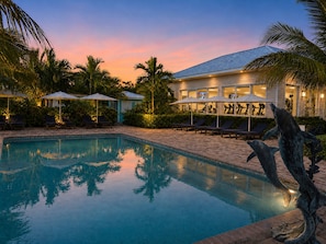 A swimming pool with lounge chairs and umbrellas, surrounded by palm trees and a building with exercise equipment visible through the windows. - Brookes Guest House (Freeport)