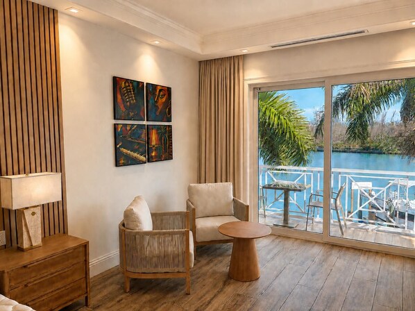 A room with a wooden floor, a sofa, a chair, a small round table, and a view of a water body through large windows.