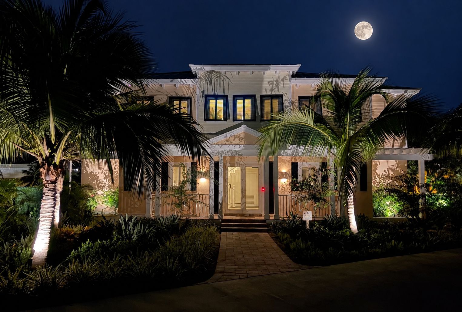 A two-story house with a well-lit entrance, surrounded by palm trees and a paved pathway.