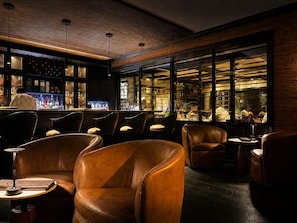A bar area with leather chairs, a bar counter, and a view of the kitchen through large windows.