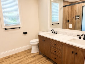 Un baño con inodoro blanco, tocador de madera con cajones, lavabo doble con grifos negros y un espejo grande.