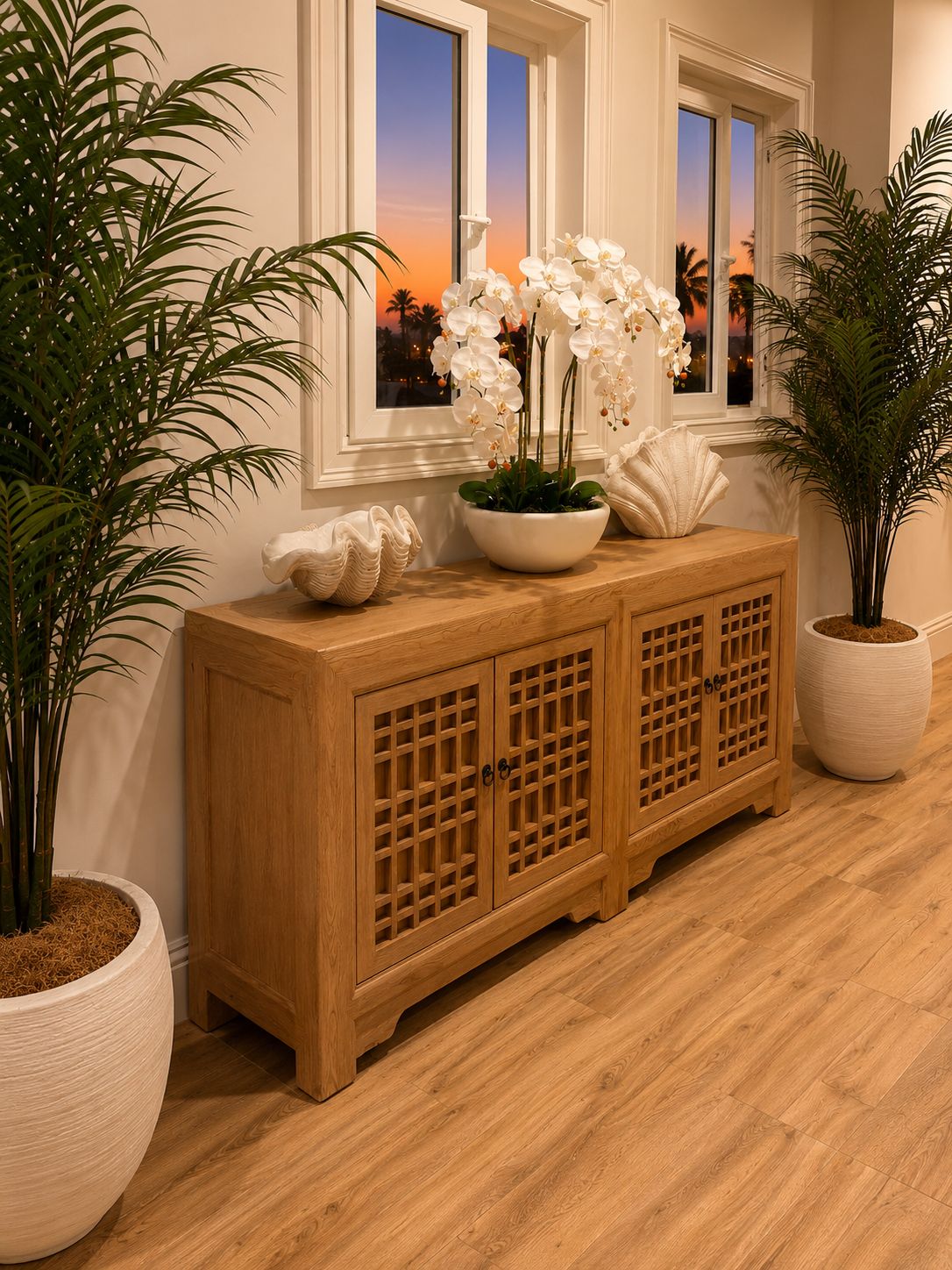 Um armário de madeira com uma tigela decorativa e orquídeas brancas, acompanhado por plantas em vasos e uma janela com vista para o pôr do sol.