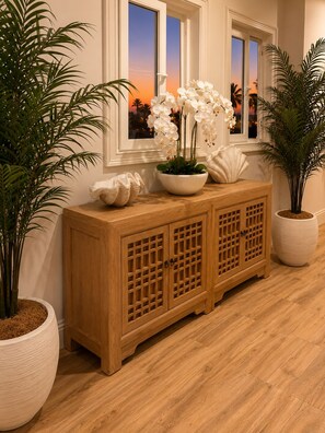A wooden cabinet with a decorative bowl and white orchids, flanked by potted plants and a window with a sunset view.