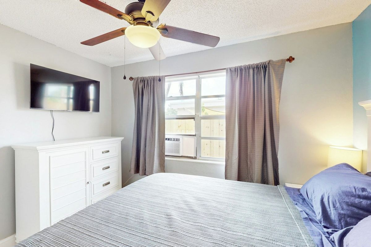 A bedroom with a bed, a ceiling fan, a TV mounted on the wall, and a window with curtains.