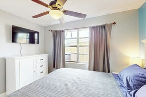 A bedroom with a bed, a ceiling fan, a TV mounted on the wall, and a window with curtains. - Scuba Shack - Enclosed Backyard (West Palm Beach)