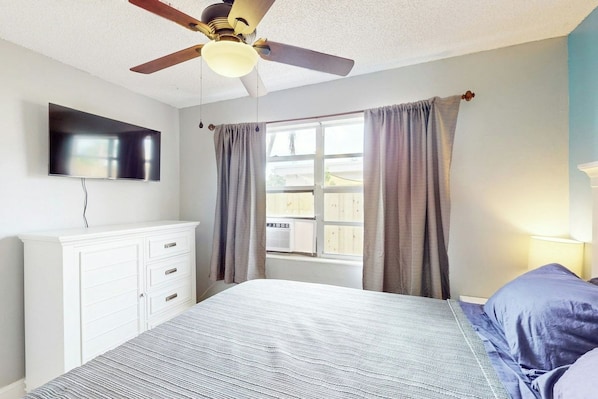 A bedroom with a bed, a ceiling fan, a TV mounted on the wall, and a window with curtains. - Scuba Shack - Enclosed Backyard (West Palm Beach)