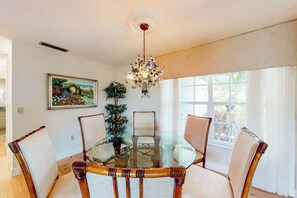 A dining area with a glass table surrounded by chairs, a chandelier, a painting, and a window with curtains. - Singer Island Serenity (West Palm Beach)
