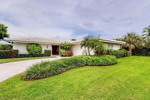A single-story house with a tiled roof, a front door, and a well-maintained lawn with various shrubs and a palm tree. - Singer Island Serenity (West Palm Beach)