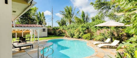 Piscina com deck de pedra, espreguiçadeiras e área de estar coberta.