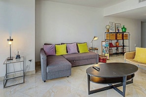 A modern living room with a grey sectional sofa, a round coffee table, and a yellow armchair. There is a small side table with a lamp and a shelf with baskets and decorative items. - Iconic Views - Luxury Condo in the Heart of Brickell (Miami)