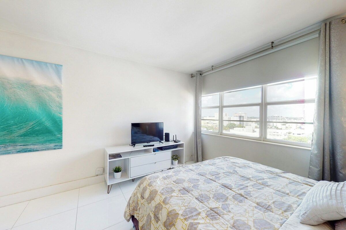 Un dormitorio con cama, un televisor sobre un mueble blanco, ventana con cortinas y un cuadro en la pared.