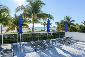 A sunlit terrace with lounge chairs and umbrellas, overlooking a beach with palm trees and a clear sky. - Decoplage Paradise - Unit A (Miami Beach)