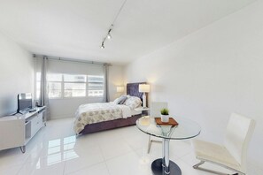 A modern hotel room with a bed, a desk, a chair, a glass table, and a window. - Decoplage Paradise - Unit A (Miami Beach)