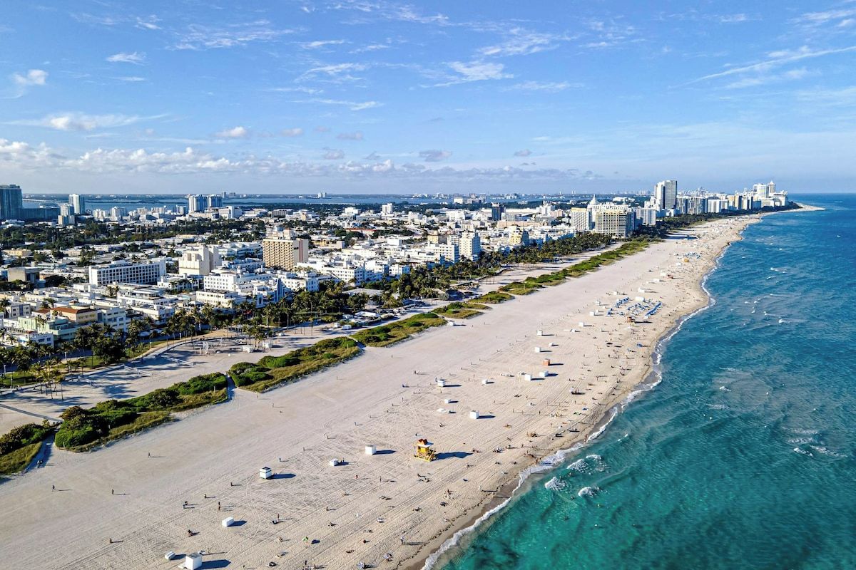 A coastal cityscape with a wide sandy beach, a clear blue sky, and a dense urban area with high-rise buildings.