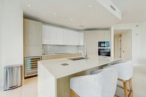 A modern kitchen with white cabinetry, a central island with bar stools, and a stainless steel trash can. - Tranquil Tides - Beachfront (Riviera Beach)