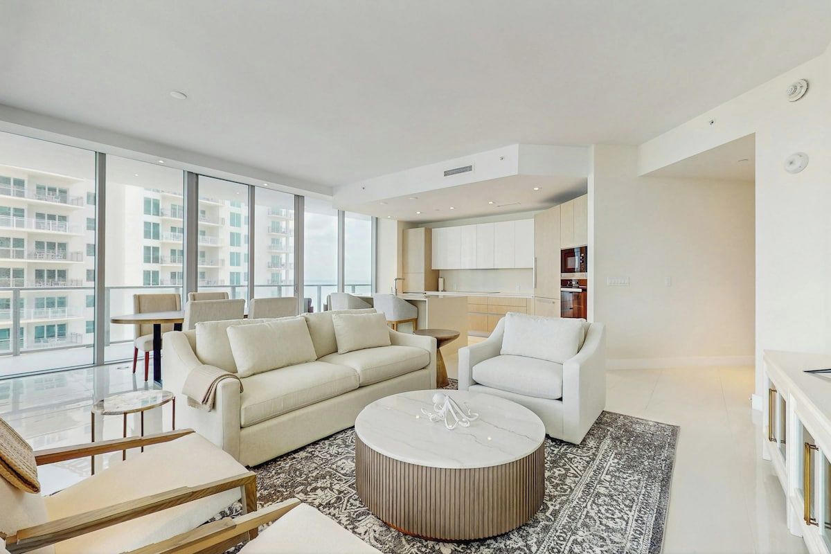 A modern living room with a sofa, armchairs, a coffee table, and a view of the city through large windows.