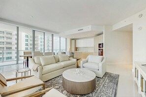 A modern living room with a sofa, armchairs, a coffee table, and a view of the city through large windows. - Tranquil Tides - Beachfront (Riviera Beach)