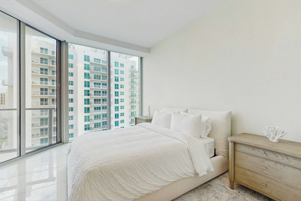 A bedroom with a large window, a bed, a nightstand, and a dresser.