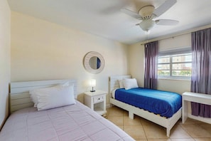 Ein Schlafzimmer mit zwei Betten, einem Deckenventilator, einem Fenster mit Vorhängen und einem Nachttisch mit Lampe.