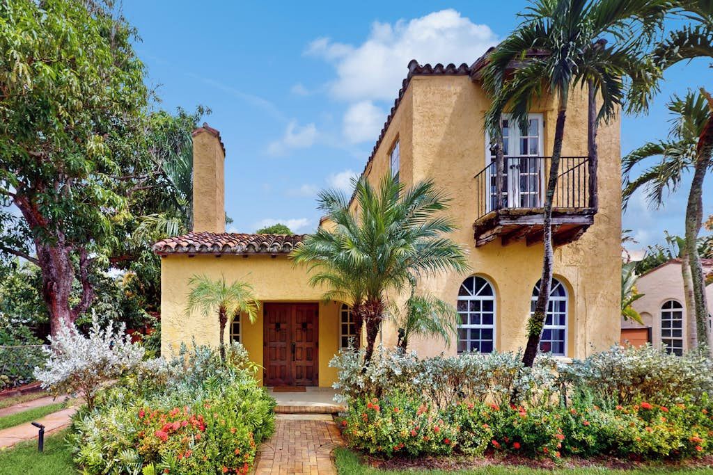 Edifício de estuque amarelo com varanda, cercado por vegetação exuberante e plantas floridas.