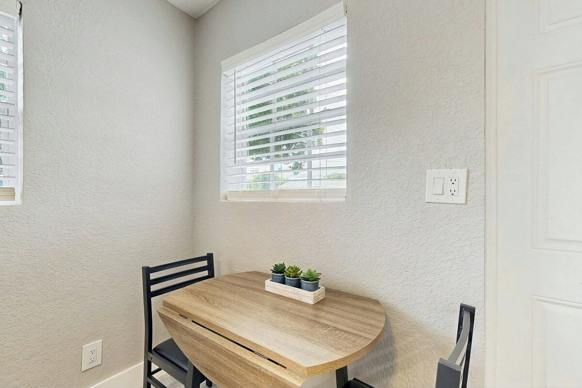 A corner room with a round wooden table, a black chair, and a window with blinds.