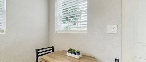 A corner room with a round wooden table, a black chair, and a window with blinds.