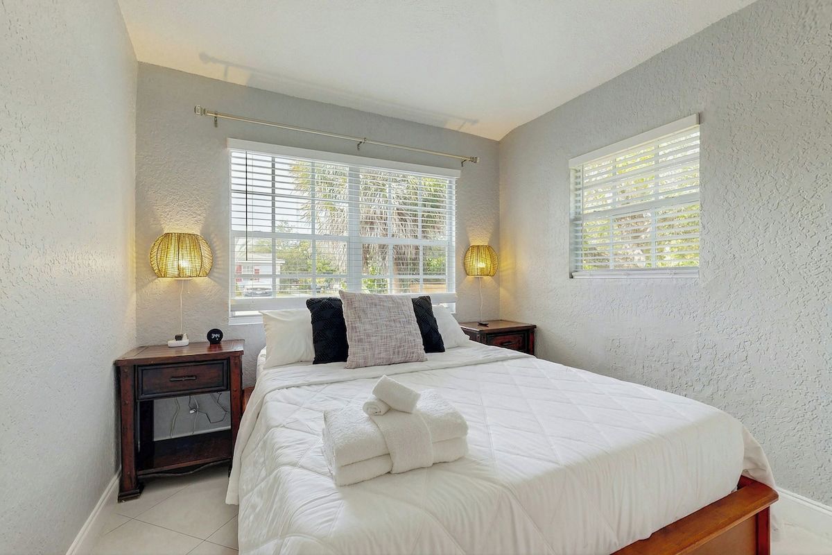 A bedroom with a large bed, two bedside tables, a window with blinds, and a wall-mounted lamp.
