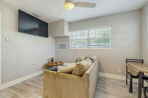 Ein Wohnzimmer mit einer Couch, einem Esstisch, einem an der Wand befestigten Fernseher und einem Deckenventilator.