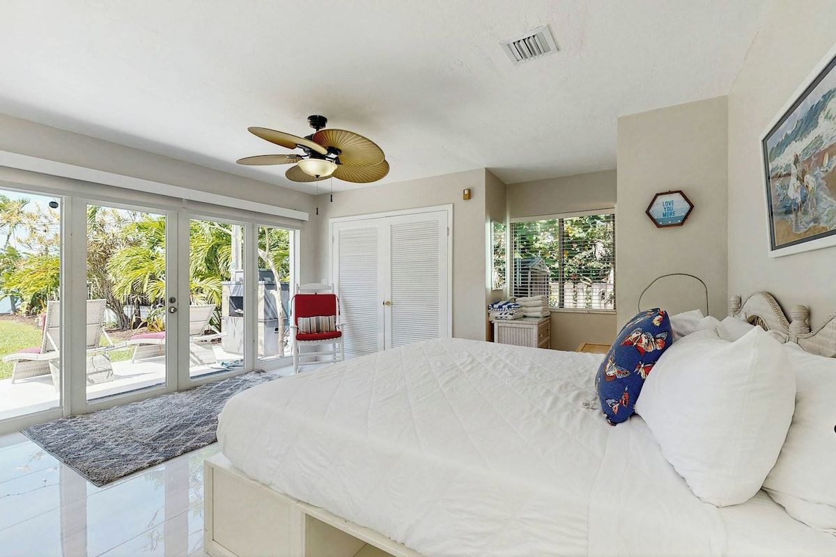 A bedroom with a large bed, a ceiling fan, a view of the outdoors, and a door leading to a patio area.